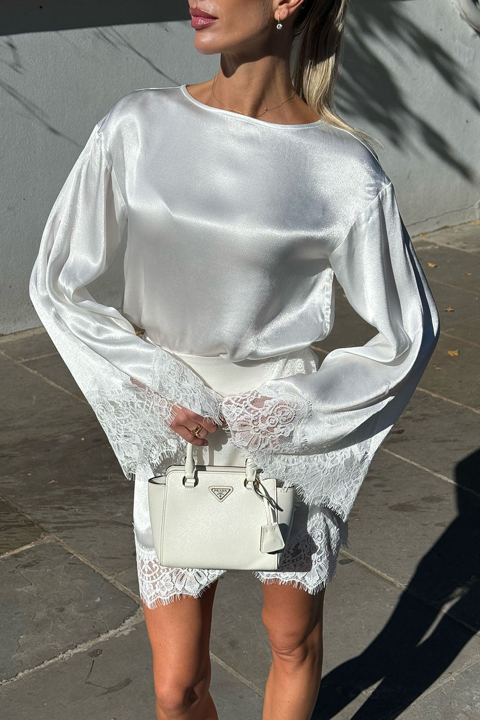 A woman wearing a white satin long-sleeve top with lace hem and holding a white Prada handbag, standing outdoors against a gray wall.