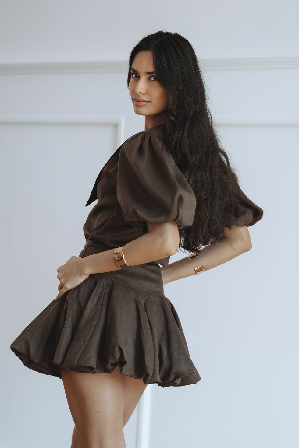 A woman modeling a dark brown puff-sleeve mini dress with a pleated skirt, posed against a clean white paneled wall.