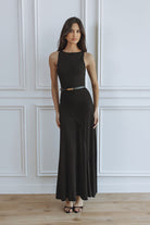 A woman modeling a sleeveless black maxi dress with a thin gold belt, standing against a white paneled wall on a light wood floor.