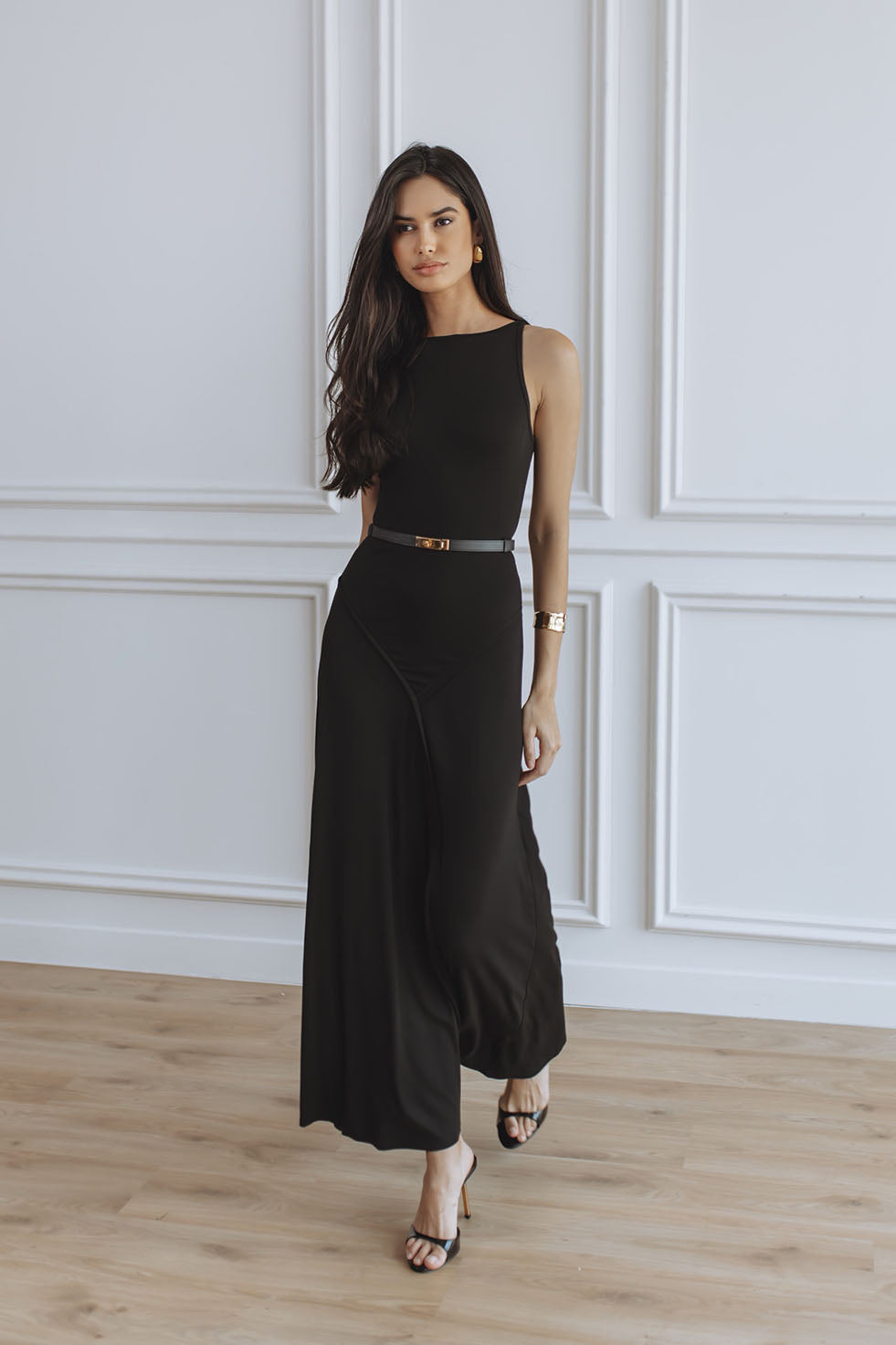 A woman modeling a black sleeveless maxi dress with a thin metallic belt at the waist, standing on a light wood floor against a white paneled wall.