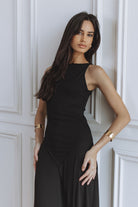 A woman wearing a sleeveless black midi dress with a draped, gathered skirt, posing against a white paneled wall.