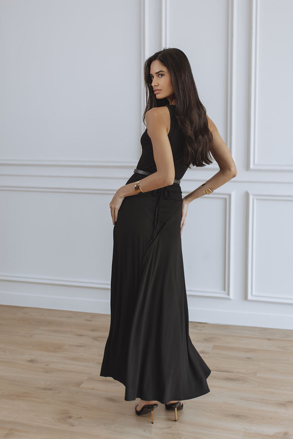 A woman modeling a black sleeveless maxi dress with a high-waisted, flared skirt in a bright room with white paneled walls and light wood floors.